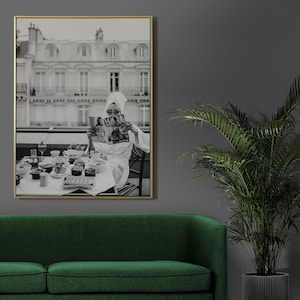 May include: Black and white photograph of a woman in a white robe sitting at a table with a magazine in her hands. The table is set with a variety of food and drinks. The woman is looking at the magazine. The photo is framed in a gold frame.