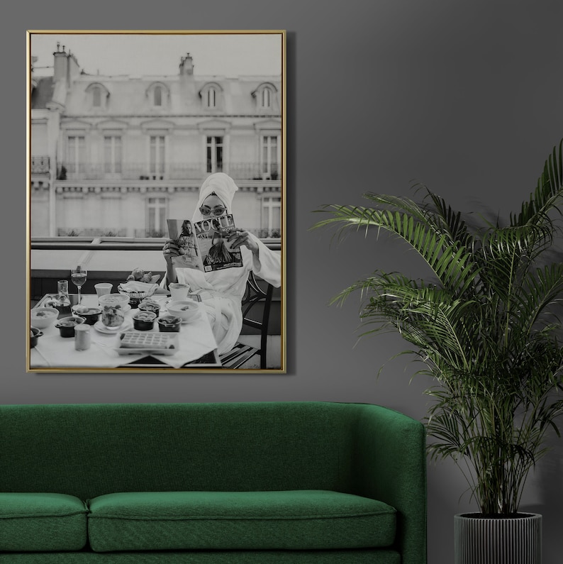 May include: Black and white photograph of a woman in a white robe sitting at a table with a magazine in her hands. The table is set with a variety of food and drinks. The woman is looking at the magazine. The photo is framed in a gold frame.