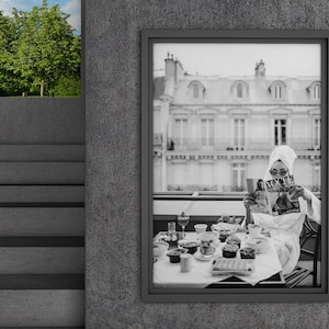 May include: Black and white photograph of a woman in a white bathrobe reading a magazine while sitting at a table with a breakfast spread. The woman is wearing sunglasses and a white towel on her head. The table is set with a variety of food and drinks, including a glass of wine, a plate of pastries, and a bowl of fruit. The photograph is framed in a black frame and is hanging on a gray wall.