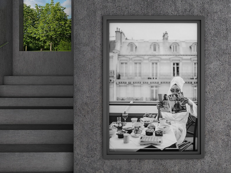 May include: Black and white photograph of a woman in a white bathrobe reading a magazine while sitting at a table with a breakfast spread. The woman is wearing sunglasses and a white towel on her head. The table is set with a variety of food and drinks, including a glass of wine, a plate of pastries, and a bowl of fruit. The photograph is framed in a black frame and is hanging on a gray wall.