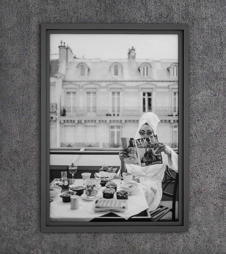 May include: A black and white photograph of a person in a white robe sitting at a table with a spread of food and reading a magazine. The magazine cover features the word "Vogue" in large letters. The person is sitting on a balcony overlooking a city.
