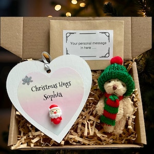 May include: A festive gift box with a heart-shaped ornament inscribed with "Christmas Hugs Sophia", a teddy bear in a green hat and scarf, and a card with the text "Your personal message in here....". The box is filled with shredded paper.