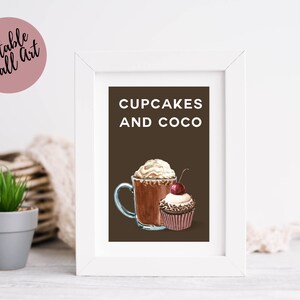 May include: White framed wall art with the words "CUPCAKES AND COCO" on a brown background. The art features a watercolor illustration of a cup of hot chocolate with whipped cream and a cupcake topped with a cherry. A pink circle with the words "Printable Wall Art" is in the upper left corner.