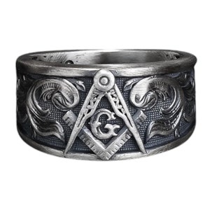Puede incluir: Un anillo de plata con un grabado detallado de una brújula y una escuadra, un símbolo común de la masonería. El anillo es ancho y tiene una superficie texturizada.