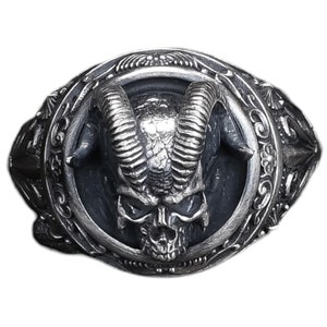 Puede incluir: Anillo plateado con un cráneo detallado y cuernos curvos. El cráneo está centrado dentro de un marco circular con adornos. El anillo tiene una estética gótica o motera.