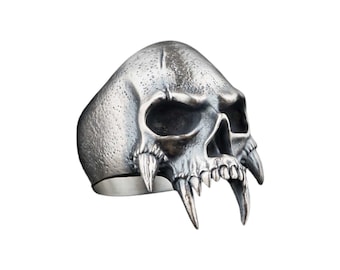 May include: A silver skull ring with large fangs. The skull is detailed with a textured surface and has a menacing expression.