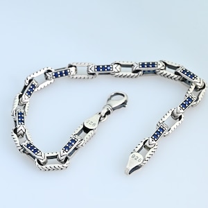 Könnte beinhalten: Ein silbernes Armband mit einem Kettenglied-Design. Jedes Glied hat eine Reihe kleiner, dunkelblauer Steine. Das Armband hat einen Karabinerverschluss. Der Verschluss und das Ende des Armbands sind mit "925" gestempelt.