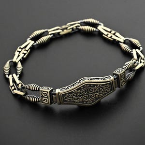 Pode incluir: Uma pulseira detalhada, de estilo antigo, com uma placa retangular central com um design floral ornamentado. A pulseira é feita de metal dourado com elos de corrente intrincados e detalhes enrolados. A pulseira é exibida contra um fundo escuro.