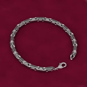 May include: A silver-toned metal chain bracelet featuring a repeating black Greek key pattern engraved on each rectangular link. The bracelet is constructed with a series of connected segments and is secured with a polished lobster claw clasp.