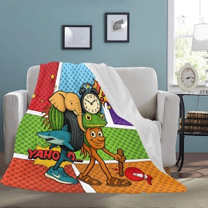 May include: A colorful comic-style blanket featuring cartoon characters including an elephant, shark, frog, and clock. The blanket has a vibrant design with the word "YAHO" in a yellow burst. The blanket is draped over a white couch.