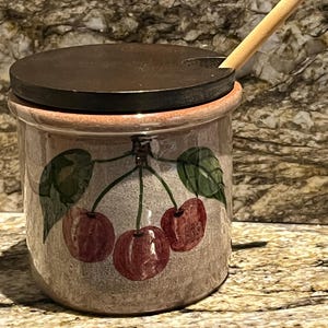 Puede incluir: Un tarro de cerámica con tapa marrón oscuro y cuchara de madera. El tarro está decorado con un diseño de cerezas en tonos rojos y verdes. El tarro tiene un borde marrón claro.