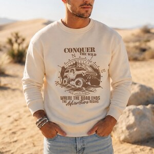 Pu&ograve; includere: Una felpa girocollo color crema con una stampa grafica marrone di un veicolo fuoristrada su uno sfondo a bussola. Il testo recita CONQUER THE WILD e WHERE THE ROAD ENDS THE ADVENTURE BEGINS in un mix di caratteri serif e corsivi.