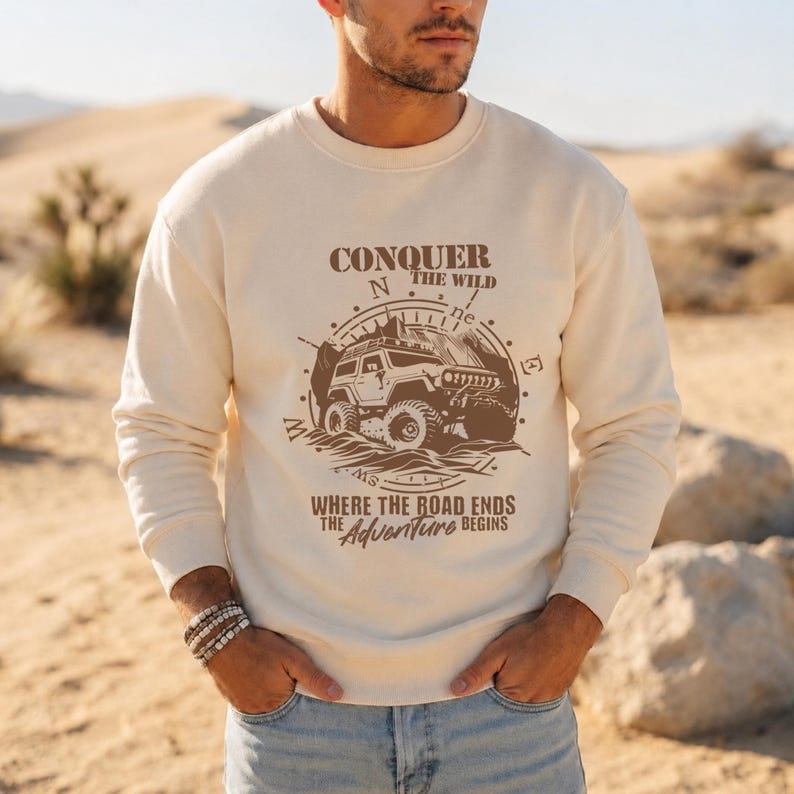 Pu&ograve; includere: Una felpa girocollo color crema con una stampa grafica marrone di un veicolo fuoristrada su uno sfondo a bussola. Il testo recita CONQUER THE WILD e WHERE THE ROAD ENDS THE ADVENTURE BEGINS in un mix di caratteri serif e corsivi.