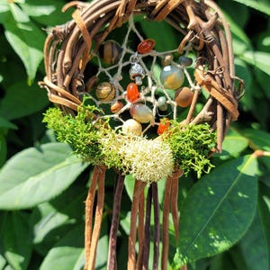 May include: A handcrafted dreamcatcher with a circular woven twig frame. The dreamcatcher features a web of twine, colorful beads, and a small mushroom charm. Green moss and leather strips hang from the bottom, set against a backdrop of green leaves.