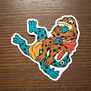 Könnte beinhalten: Ein Aufkleber mit zwei stilisierten Fröschen in einer Wrestling-Pose. Ein Frosch ist braun mit schwarzen Punkten und einem roten Schal, der andere ist blau mit schwarzen Punkten. Auf weißem Hintergrund.
