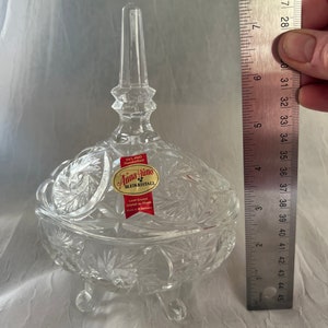 May include: A clear, lead crystal candy dish with a lid, resting on three small feet. The dish features a starburst pattern and a tall, pointed finial. The label reads "Anna Hütte Bleikristall". The dish measures approximately 10 cm in height.
