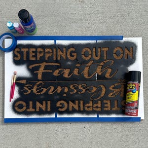 May include: A sign in progress with the words "STEPPING OUT ON Faith & Blessings" stenciled in brown on a black background. Blue tape, paint bottles, and a brush are visible.