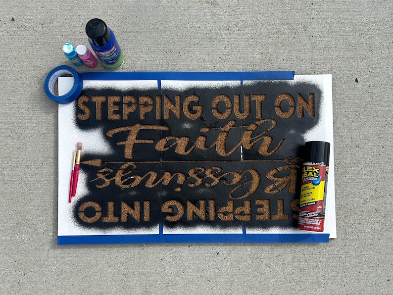 May include: A sign in progress with the words "STEPPING OUT ON Faith & Blessings" stenciled in brown on a black background. Blue tape, paint bottles, and a brush are visible.