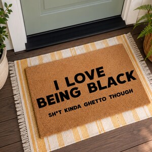 May include: A rectangular doormat made of natural coir fiber, with the text "I LOVE BEING BLACK SH*T KINDA GHETTO THOUGH" in bold black letters. The mat sits on a yellow and white striped rug.
