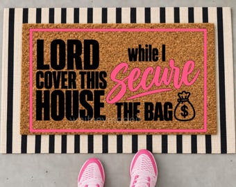 May include: A rectangular tan coir doormat with a pink border featuring the black text LORD COVER THIS HOUSE while I Secure THE BAG and a black money bag icon with a dollar sign. The mat is layered over a black and white striped rug.