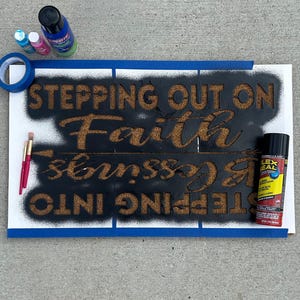 May include: A sign with the words "STEPPING OUT ON Faith INTO blessings" stenciled in brown on a black background. The sign is taped to a white surface with blue painter's tape. Paint supplies, including spray paint and paintbrushes, are visible.