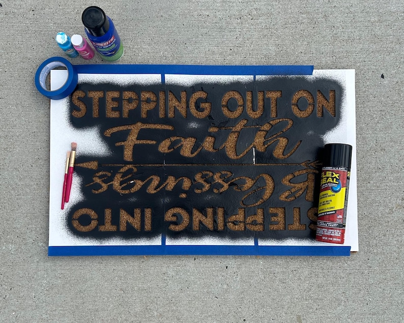 May include: A sign with the words "STEPPING OUT ON Faith INTO blessings" stenciled in brown on a black background. The sign is taped to a white surface with blue painter's tape. Paint supplies, including spray paint and paintbrushes, are visible.