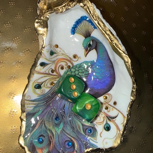 May include: A decorative oyster shell featuring a colorful peacock illustration with gold-painted edges. The shell is adorned with small embedded crystals and two green dice with gold pips resting on the surface of the peacock's tail feathers.