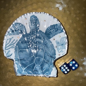 May include: A white scallop shell dish featuring a blue sea turtle illustration, accented with small embedded crystals and a silver-painted edge. Two blue dice with white pips are positioned next to the shell, which has a glossy finish.
