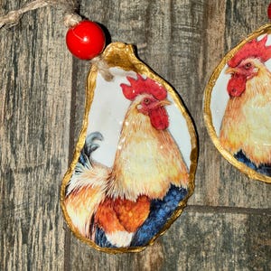 May include: Two oyster shell ornaments featuring a detailed illustration of a rooster with a bright red comb and wattles, golden-yellow neck feathers, and dark blue tail feathers. The edges are painted gold, and each includes a red bead and twine.