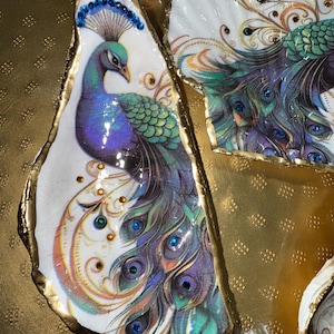 May include: Two decorative oyster and scallop shells featuring detailed peacock illustrations in shades of blue, green, and purple. The shells have gold-painted edges and are embellished with small, colorful adhesive gems on the peacock feathers.