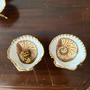 May include: Two white scallop shells with gold-painted edges, each featuring a detailed illustration of a spiral seashell in shades of brown and tan. One shell contains two translucent amber-colored dice with yellow pips resting inside.