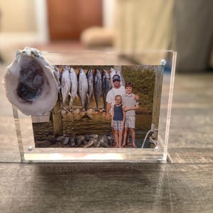 May include: Clear acrylic photo frame with a family photo and an oyster shell attached. The photo shows a group of people with fish hanging in the background. The frame is on a wooden surface.