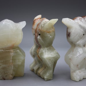May include: Three carved owl figurines made of translucent stone. The owls have a light green and white color with brown and orange veining. Each owl has pointed ears and a simple, stylized form. The background is a solid gray.