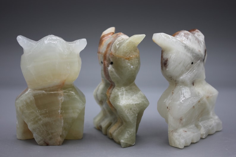 May include: Three carved owl figurines made of translucent stone. The owls have a light green and white color with brown and orange veining. Each owl has pointed ears and a simple, stylized form. The background is a solid gray.