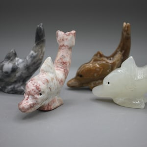 May include: Four carved stone dolphin figurines in various colors. One is gray with white marbling, another is white with red veining, a third is brown, and the last is a translucent white. Each dolphin has a small, dark eye.