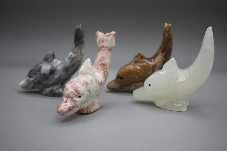 May include: Four carved stone dolphin figurines in various colors. One is gray with white marbling, another is white with red veining, a third is brown, and the last is a translucent white. Each dolphin has a small, dark eye.