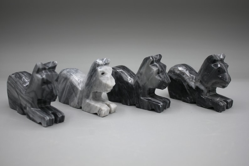 May include: Four carved stone lion sculptures in varying shades of gray and black. Each lion is in a seated position with its front paws extended. The sculptures have a geometric, angular design.