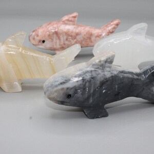 May include: Four carved stone shark figurines in shades of white, pink, and gray. Each shark has a detailed fin and tail design, with small black eyes. The sharks are displayed on a light gray surface.