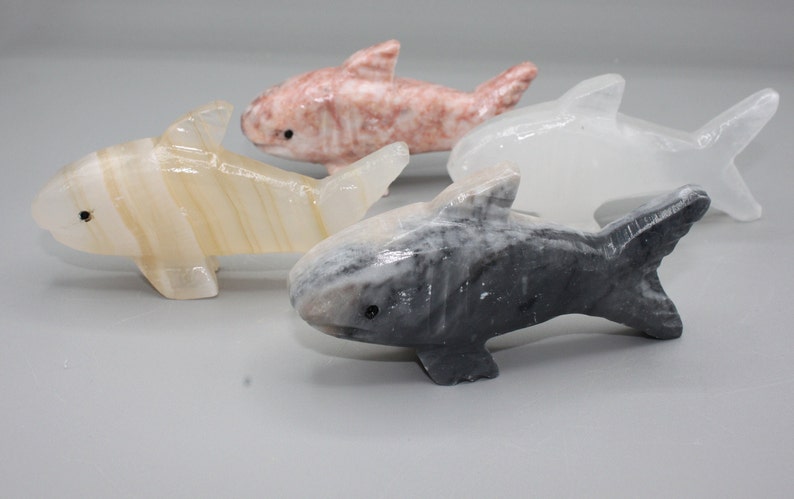 May include: Four carved stone shark figurines in shades of white, pink, and gray. Each shark has a detailed fin and tail design, with small black eyes. The sharks are displayed on a light gray surface.