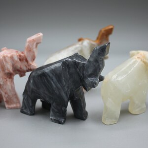 May include: A collection of carved stone elephant figurines in various colors, including pink, black, white, and brown. The elephants are stylized with geometric shapes and stand on four legs.