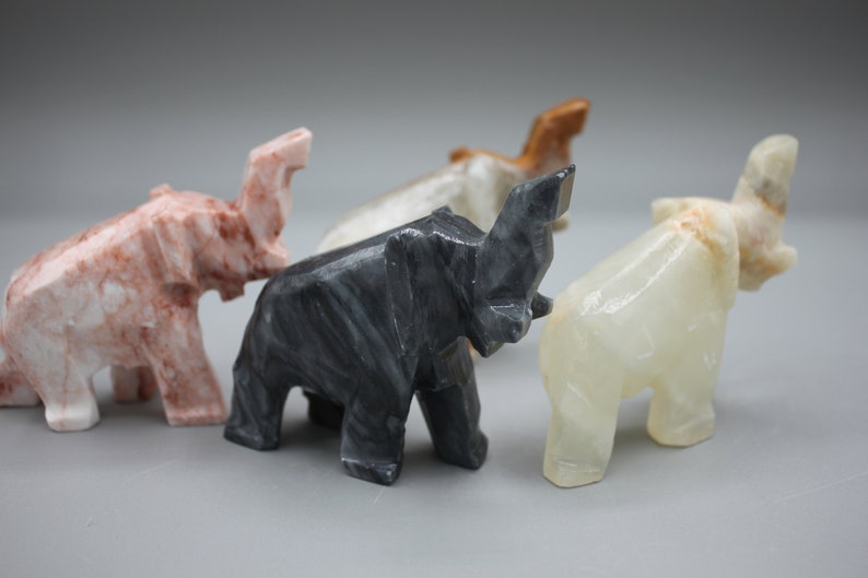 May include: A collection of carved stone elephant figurines in various colors, including pink, black, white, and brown. The elephants are stylized with geometric shapes and stand on four legs.