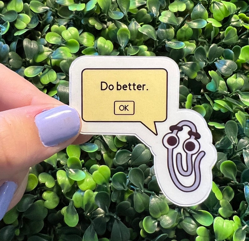 Op de afbeelding: Een witte sticker met een gele spraakballon die "Do better." zegt en een "OK"-knop. De spraakballon is bevestigd aan een grijze paperclip met ogen.