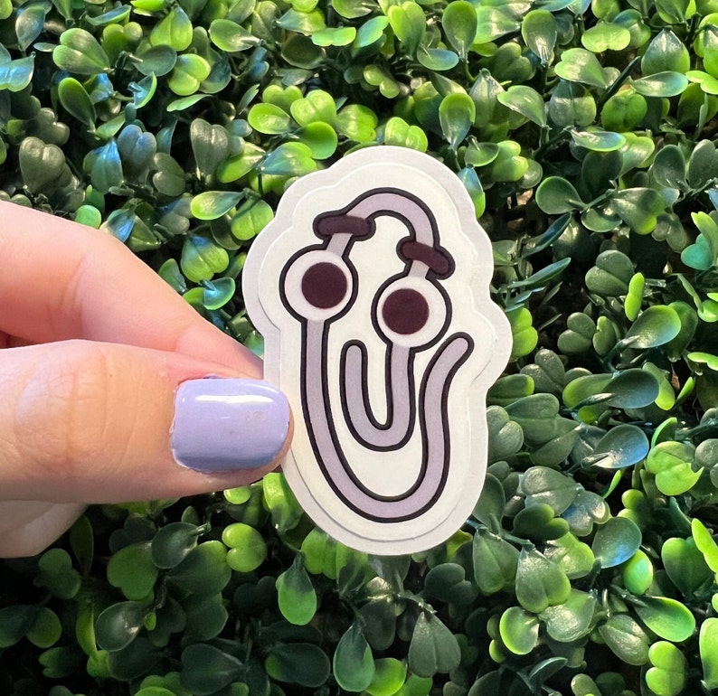Op de afbeelding: Een witte sticker met een paarse omtrek van een koptelefoon met twee grote, donkerpaarse ogen. De sticker zit op een groene, bladerige achtergrond.
