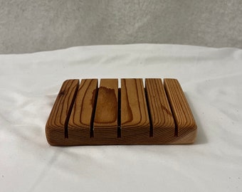May include: A rectangular wooden soap dish with a slatted design. The dish is made of light brown wood and has a natural grain pattern. The soap dish is designed to allow water to drain, extending the life of the soap.