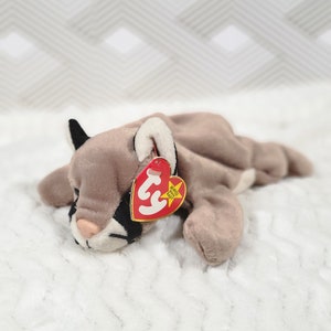 Puede incluir: Un animal de peluche marrón y blanco que se asemeja a un puma, acostado de lado con la cabeza apoyada en las patas. El animal de peluche tiene una etiqueta Ty roja con una estrella blanca y el texto "Beanie Baby" en ella.