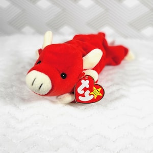 Puede incluir: Toro de peluche rojo Beanie Baby con cuernos blancos y una cara blanca. El Beanie Baby tiene una etiqueta blanca con las palabras "Ty" y "Beanie Original Baby" en amarillo.