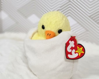 Peut inclure: Un poussin en peluche jaune dans un œuf en peluche blanc. Le poussin a un bec rouge et des yeux noirs. L'œuf a une étiquette Ty Beanie Babies rouge et blanche attachée.