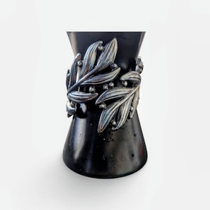 Könnte beinhalten: Ein silberfarbenes Armband mit einem detaillierten Reliefdesign aus überlappenden Blättern und kleinen kugelförmigen Akzenten. Das Metallstück ist um einen glatten, mattschwarzen konischen Ständer gewickelt, der die komplexe botanische Textur hervorhebt.