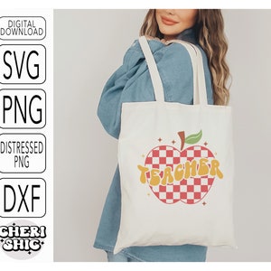 May include: A white tote bag with a red and white checkered apple design and the word "TEACHER" in yellow letters. The bag is being held by a person wearing a blue denim shirt.