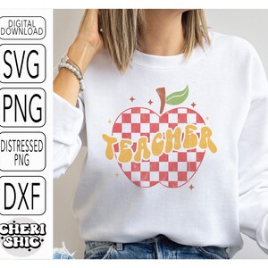 May include: White sweatshirt with a red and white checkered apple design and the word "TEACHER" in yellow letters.